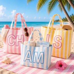 May include: Three canvas tote bags with vertical stripes in pink, blue, and yellow. The pink bag features the letters CH, the blue bag features AM, and the yellow bag features IS, all in embroidered block lettering. Each bag has two top handles.