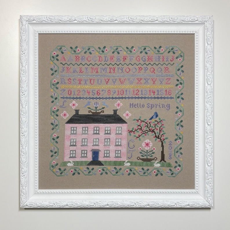 Pode incluir: Um sampler de ponto cruz emoldurado com uma casa rosa, uma &aacute;rvore com um p&aacute;ssaro azul e o texto "Hello Spring". O sampler inclui um alfabeto, n&uacute;meros e uma borda floral. A moldura &eacute; branca.