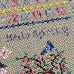 Pode incluir: Bordado em ponto cruz com as palavras "Hello Spring" em roxo. O desenho inclui um p&aacute;ssaro azul empoleirado num galho florido com flores cor-de-rosa e folhas verdes. N&uacute;meros e o alfabeto tamb&eacute;m s&atilde;o bordados.