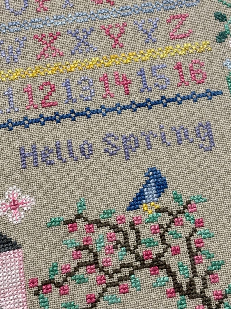 Pode incluir: Bordado em ponto cruz com as palavras "Hello Spring" em roxo. O desenho inclui um p&aacute;ssaro azul empoleirado num galho florido com flores cor-de-rosa e folhas verdes. N&uacute;meros e o alfabeto tamb&eacute;m s&atilde;o bordados.