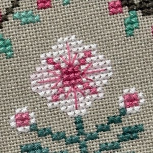 Pode incluir: Close-up de um design floral em ponto cruz. A flor central &eacute; branca com um centro rosa e linhas rosa radiantes. Duas pequenas flores rosa e brancas ladeiam a flor principal. Folhas e caules verdes tamb&eacute;m s&atilde;o vis&iacute;veis.