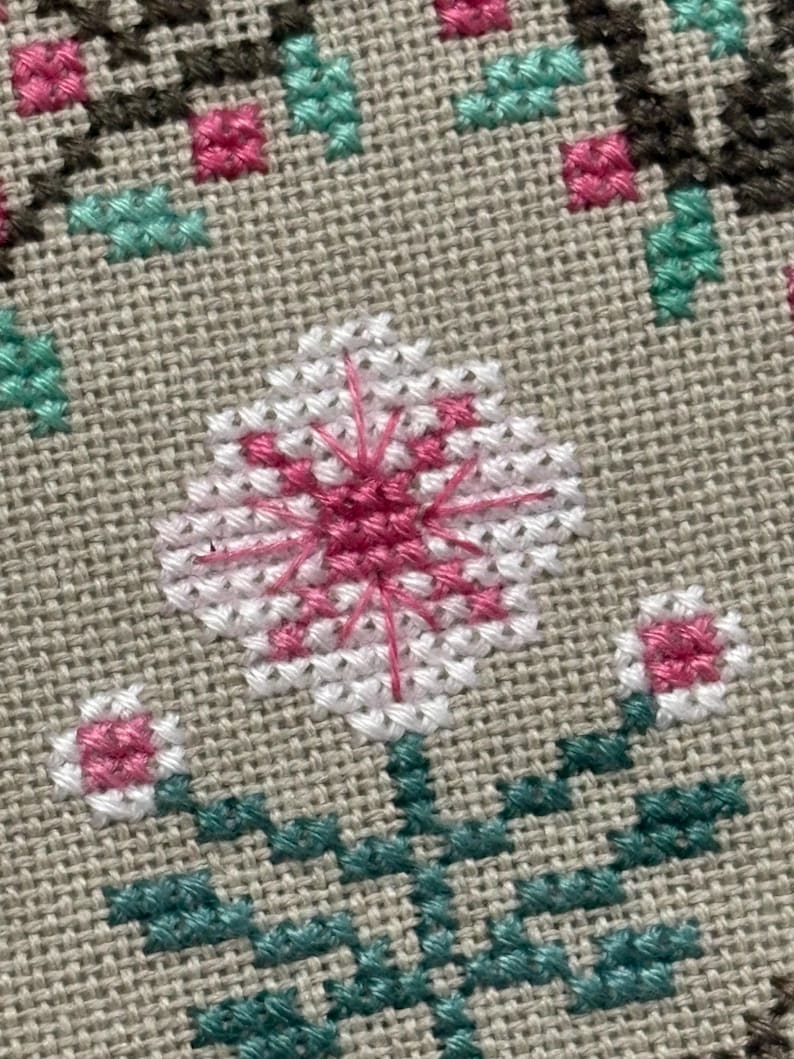 Pode incluir: Close-up de um design floral em ponto cruz. A flor central &eacute; branca com um centro rosa e linhas rosa radiantes. Duas pequenas flores rosa e brancas ladeiam a flor principal. Folhas e caules verdes tamb&eacute;m s&atilde;o vis&iacute;veis.