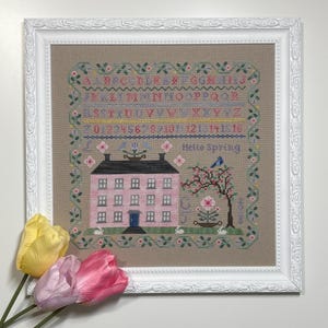 Pode incluir: Amostra de ponto cruz emoldurada com uma casa rosa, borda floral e o texto "Hello Spring". A amostra inclui um alfabeto, n&uacute;meros e elementos decorativos. A moldura &eacute; branca com um design floral detalhado. Tr&ecirc;s tulipas em tons de amarelo e rosa em primeiro plano.