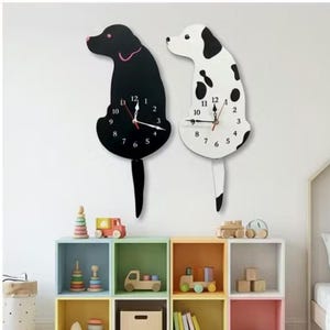Puede incluir: Dos relojes de pared con forma de perro. Uno es negro con un collar rosa, el otro es blanco con manchas negras. Ambos tienen colas negras y esferas con números y manecillas negras. Cuelgan de una pared blanca.