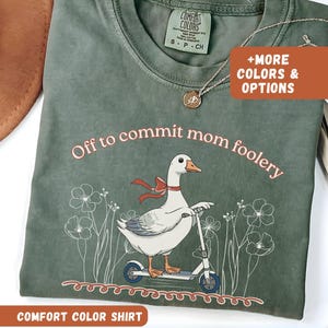 May include: A sage green Comfort Colors shirt with the phrase "Off to commit mom foolery" and a graphic of a goose on a scooter. The shirt also has the text "COMFORT COLOR SHIRT". A necklace is draped on the shirt.