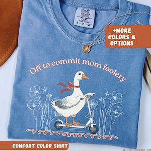 May include: A blue Comfort Colors t-shirt with the phrase "Off to commit mom foolery" and a graphic of a goose on a scooter. The shirt has a crew neck and short sleeves. A gold necklace is draped on the shirt.