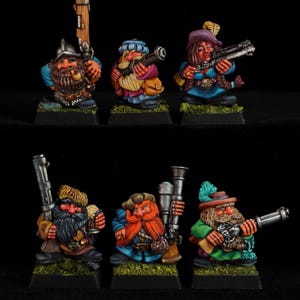 Op de afbeelding: Zes handbeschilderde miniatuur dwergfiguurtjes op vierkante zwarte voetstukken met groene, getextureerde bovenkanten. Elk figuur draagt gedetailleerde historische kleding en draagt een uniek vuurwapen, waaronder musketten en donderbussen.