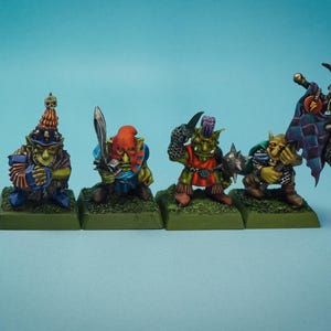 Könnte beinhalten: Vier Miniatur-Fantasy-Figuren auf grünen Sockeln. Die Figuren sind in verschiedenen Farben bemalt, darunter Grün, Blau, Rot und Braun. Eine Figur hält ein Schwert, eine andere einen Schild und eine weitere ein Banner. Die Figuren stehen vor einem hellblauen Hintergrund.