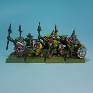 May include: A collection of miniature fantasy figures, possibly for tabletop gaming. The green-skinned figures have red eyes, wear armour and helmets, and wield spears and shields. The shields display different designs, and the figures are positioned on green bases.