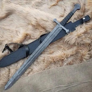 Puede incluir: Una espada larga y gris con una hoja estampada y una guarda en forma de cruz descansa en una vaina de cuero negro. El mango est&aacute; envuelto en material negro. La espada y la vaina se exhiben sobre una superficie texturizada de color marr&oacute;n claro.
