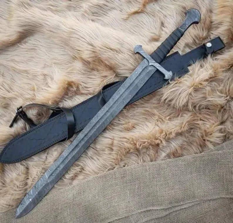Puede incluir: Una espada larga y gris con una hoja estampada y una guarda en forma de cruz descansa en una vaina de cuero negro. El mango est&aacute; envuelto en material negro. La espada y la vaina se exhiben sobre una superficie texturizada de color marr&oacute;n claro.