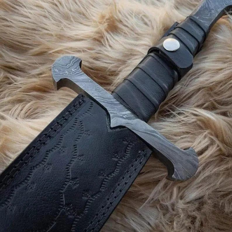 Puede incluir: Primer plano de una espada de estilo medieval con una hoja de acero estampada y un mango envuelto en cuero negro. La espada est&aacute; parcialmente enfundada en una vaina de cuero negro con costuras decorativas. El fondo es una superficie texturizada de color marr&oacute;n claro.