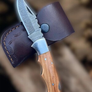 Puede incluir: Un cuchillo plegable con una hoja de acero estampada y un mango de madera. El cuchillo est&aacute; parcialmente abierto, revelando el intrincado dise&ntilde;o de la hoja. Descansa en una funda de cuero marr&oacute;n con cierre de bot&oacute;n a presi&oacute;n.
