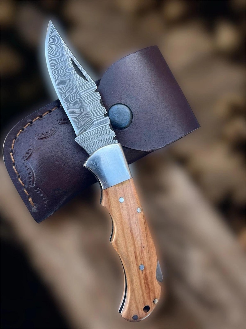 Puede incluir: Un cuchillo plegable con una hoja de acero estampada y un mango de madera. El cuchillo est&aacute; parcialmente abierto, revelando el intrincado dise&ntilde;o de la hoja. Descansa en una funda de cuero marr&oacute;n con cierre de bot&oacute;n a presi&oacute;n.