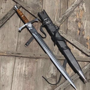 May include: A steel sword with a polished blade, a crossguard, and a brown wood-grain handle with metallic accents. Beside the sword lies a black leather scabbard featuring integrated black leather straps and a pointed metallic tip at the end.