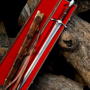Puede incluir: Una espada larga y plateada con empu&ntilde;adura y guarda de madera descansa en una caja de madera forrada en rojo. Tambi&eacute;n hay una vaina de cuero a juego con grabados decorativos y una correa. La espada y la vaina est&aacute;n sobre una superficie de madera r&uacute;stica.