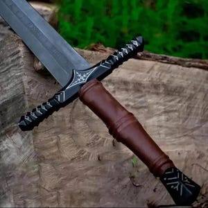 Puede incluir: Una espada negra y plateada con un mango envuelto en cuero marrón. La hoja es de color gris oscuro con detalles grabados. La empuñadura presenta intrincados diseños plateados. La espada descansa sobre una superficie de madera texturizada.