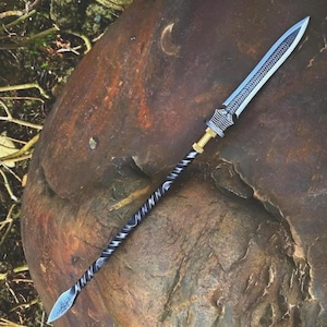 May include: A handcrafted spear with a silver blade and a black and silver twisted handle. The spearhead has a decorative pattern. The handle is connected to a gold-coloured metal piece. The spear is lying on a brown stone surface.