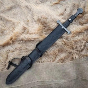 Puede incluir: Una espada de estilo medieval con una hoja plateada y un mango envuelto en cuero negro. La espada est&aacute; enfundada en una vaina de cuero negro con una trabilla para el cintur&oacute;n. El fondo es una superficie texturizada de color marr&oacute;n claro.