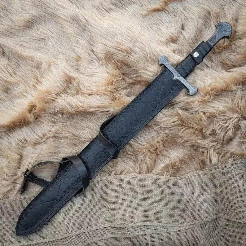 Puede incluir: Una espada de estilo medieval con una hoja plateada y un mango envuelto en cuero negro. La espada est&aacute; enfundada en una vaina de cuero negro con una trabilla para el cintur&oacute;n. El fondo es una superficie texturizada de color marr&oacute;n claro.