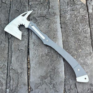 Puede incluir: Un hacha táctica plateada y gris con un mango curvo. La cabeza del hacha tiene un diseño único con una hoja curva, una parte superior puntiaguda y un recorte circular. El mango es gris con detalles plateados.