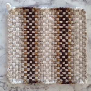 May include: Square, woven potholder in neutral tones. The design features a repeating pattern of light beige, brown, and dark brown squares. Two vertical stripes of dark brown squares run down the center. A small loop is attached to one corner.