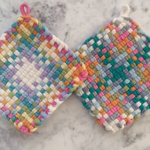 May include: Two woven potholders in a square shape. One has a pink loop, the other a white loop. The potholders are made of colorful woven fabric in shades of pink, blue, green, yellow, and white. The weave pattern is visible.
