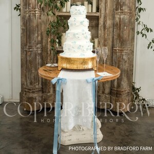 May include: A five-tier white wedding cake decorated with light blue floral accents and swags, resting on a gold pedestal stand. The cake sits on a round wooden table draped with a white linen runner featuring light blue ribbon ties.