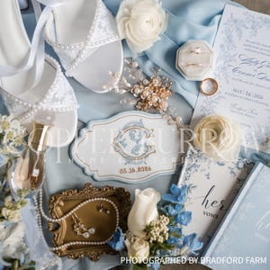 May include: A wedding flat lay featuring white pearl-embellished heels, a gold-toned hair accessory, a ring box with two rings, a blue and white invitation, a custom embroidered patch with initials and 05.16.2026, and a vow book.