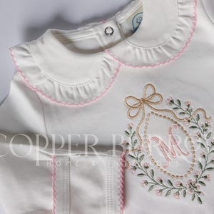 Könnte beinhalten: Ein weißes Langarmshirt mit einem gerüschten Kragen mit rosa Saum, einem Metalldruckknopf am Hals und einem gestickten ovalen Kranzdesign mit einem zentralen Buchstaben M und einer Schleife. Der Text COPPER BURROW HOME & INTERIORS ist sichtbar.