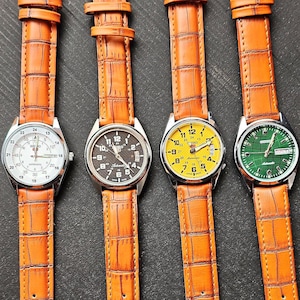May include: Four wristwatches with orange leather straps and silver watch faces. The watch faces are white, black, yellow, and green. The watch straps have a crocodile skin texture.
