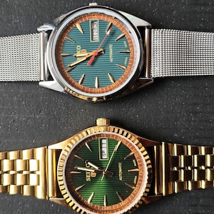Peut inclure: Deux montres-bracelets avec des cadrans verts et des bracelets métalliques. La montre du haut a un bracelet en maille argentée, celle du bas un bracelet à maillons dorés. Les deux montres affichent la date et ont des détails dorés.