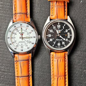 May include: Two wristwatches with stainless steel cases and orange leather straps. One watch has a white face with black numerals, the other has a black face with white numerals. Both watches have a date display.