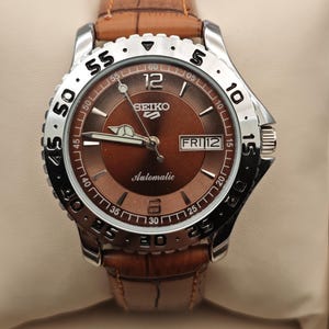 May include: A Seiko 5 automatic wristwatch with a brown sunray dial, silver-tone hands, and indices. The watch features a stainless steel bezel with black numerals, a day-date window at the three o'clock position, and a brown leather strap.