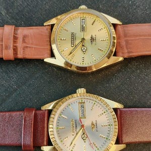 May include: Two Citizen automatic wristwatches with gold-toned cases and brown leather straps. The watch faces are gold and silver, displaying the day and date. The watches have a classic design.