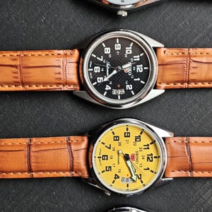 May include: Four wristwatches with silver cases and brown leather straps are displayed. The watch faces are white, black, yellow, and green. Each watch has a different colored face with black numerals and hands. The watch straps have a textured pattern.