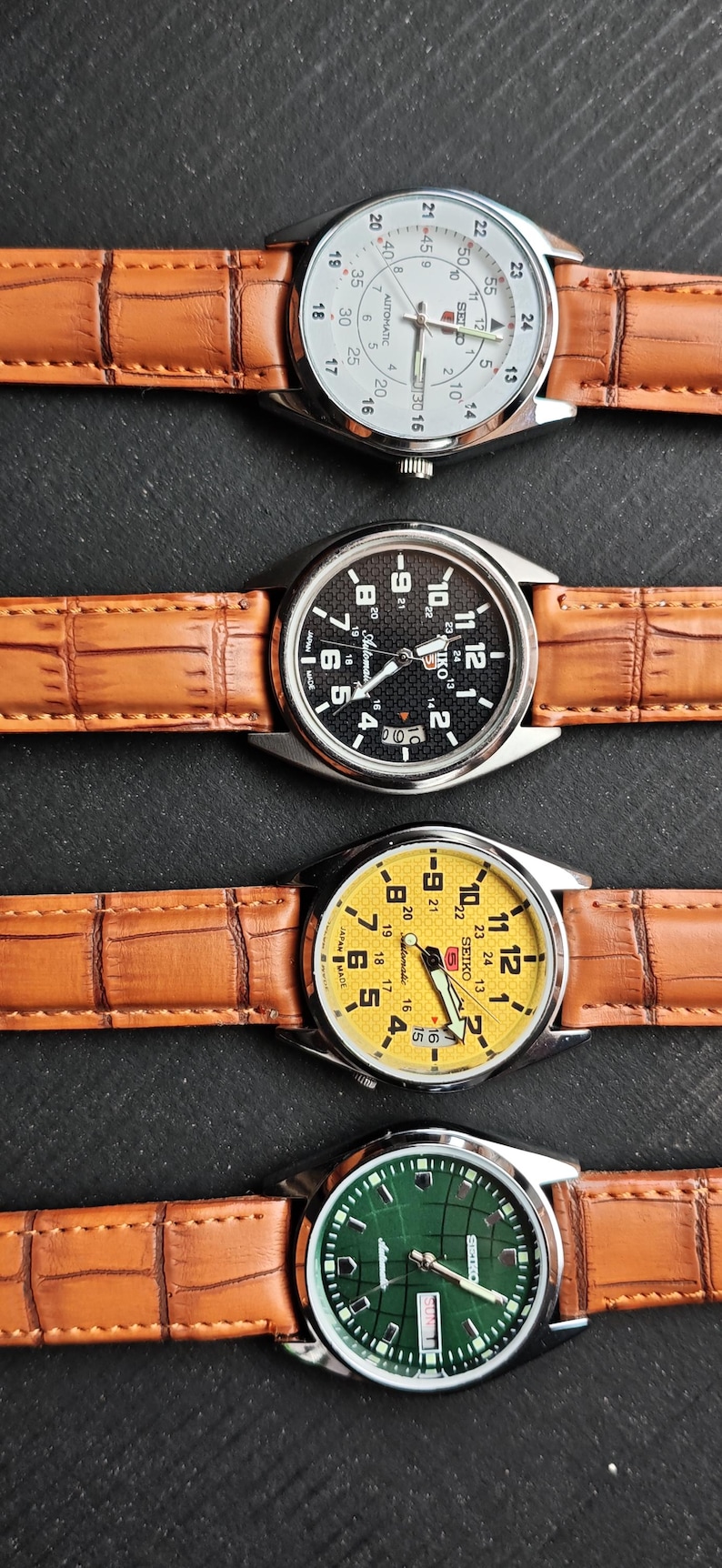 May include: Four wristwatches with silver cases and brown leather straps are displayed. The watch faces are white, black, yellow, and green. Each watch has a different colored face with black numerals and hands. The watch straps have a textured pattern.