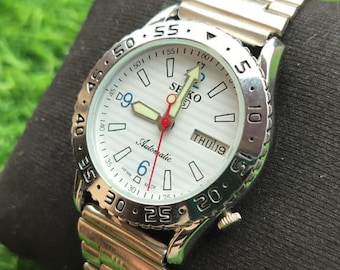 Reloj de pulsera vintage Seiko 5 con esfera blanca y bisel fijo, fecha y día automático.