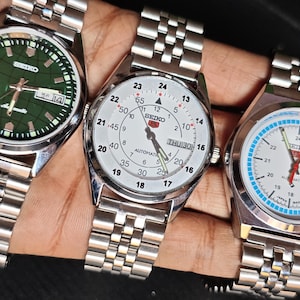 May include: Three Seiko wristwatches with stainless steel bands. The watches have different colored faces: green, white, and white with blue and orange accents. Each watch has a date display and a silver-colored case.