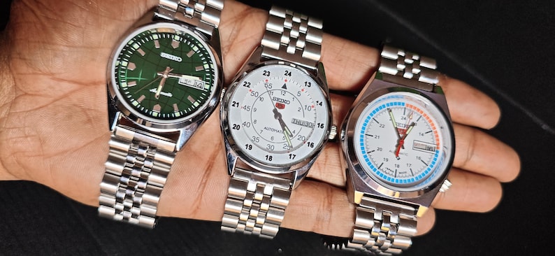 May include: Three Seiko wristwatches with stainless steel bands. The watches have different colored faces: green, white, and white with blue and orange accents. Each watch has a date display and a silver-colored case.