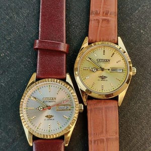 May include: Two Citizen automatic wristwatches with gold-toned cases and leather bands. One watch has a burgundy band, the other a brown, textured band. Both watches display the day and date.
