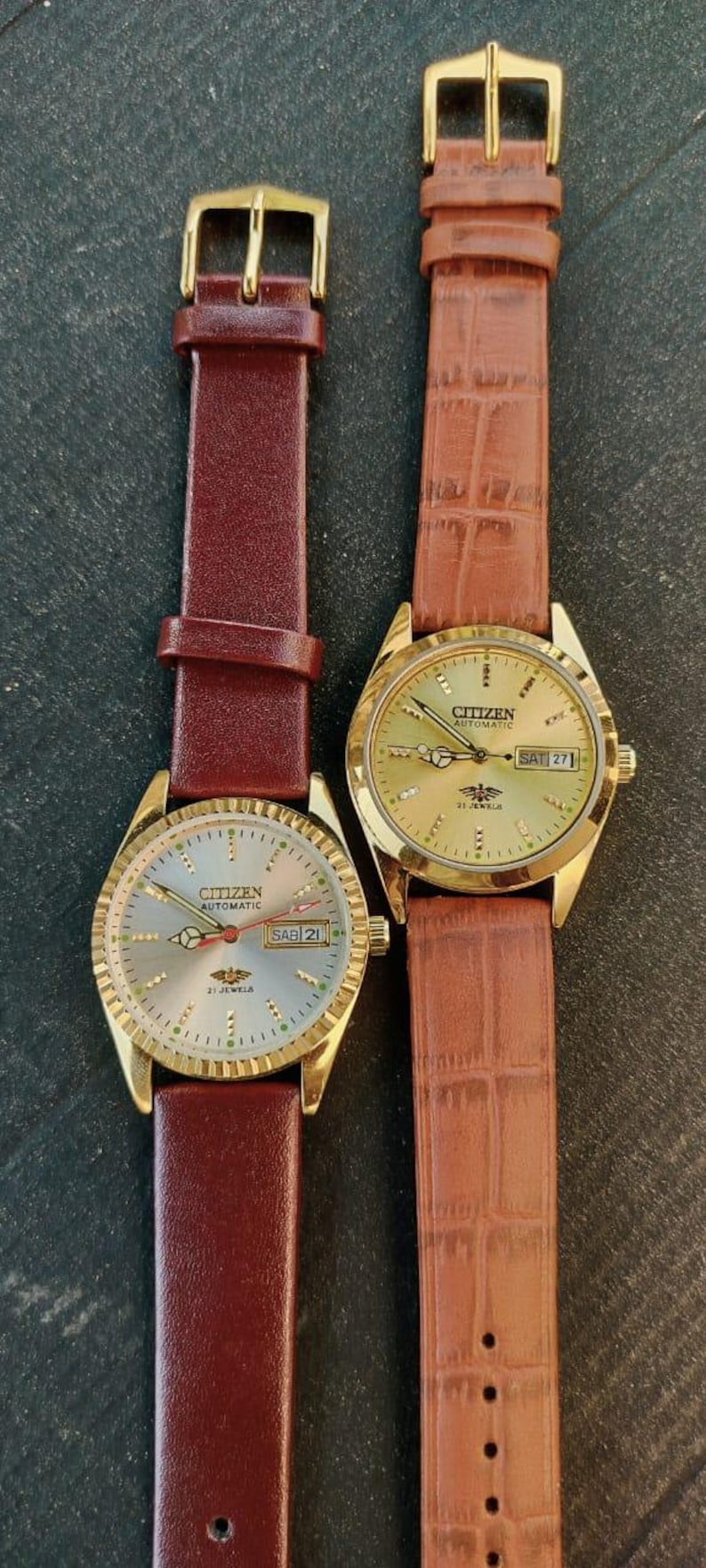 May include: Two Citizen automatic wristwatches with gold-toned cases and leather bands. One watch has a burgundy band, the other a brown, textured band. Both watches display the day and date.