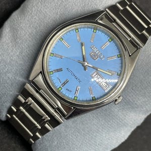 May include: A stainless steel Seiko 5 automatic wristwatch featuring a light blue dial, silver-tone baton indices with green accents, silver hands, and a day-date window at the three o'clock position. The watch includes a metal link bracelet.
