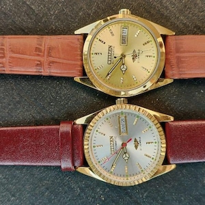 May include: Two gold-toned Citizen watches with leather straps. One watch has a brown, textured strap, and the other has a burgundy strap. Both watches have gold faces with day and date displays. The watches are displayed on a dark surface.
