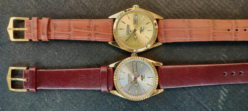 May include: Two gold-toned Citizen watches with leather straps. One watch has a brown, textured strap, and the other has a burgundy strap. Both watches have gold faces with day and date displays. The watches are displayed on a dark surface.