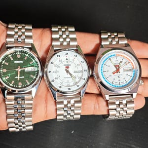 May include: Three silver-toned Seiko wristwatches with metal bands are displayed. The watches have white, green, and blue faces, with visible date displays. The watch faces have black and white markings.