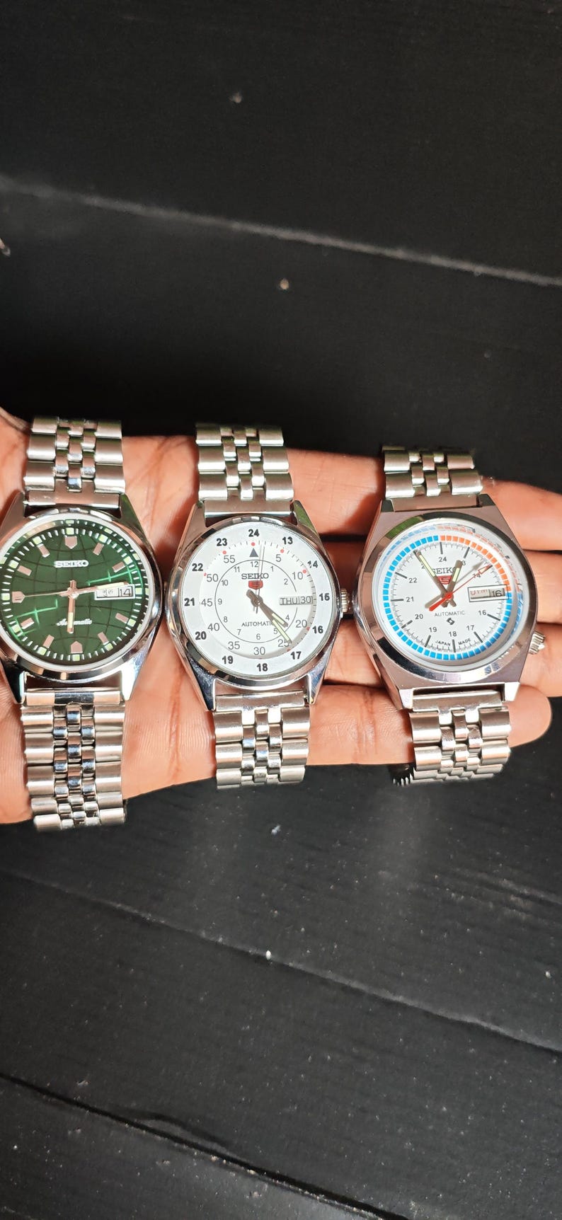 May include: Three silver-toned Seiko wristwatches with metal bands are displayed. The watches have white, green, and blue faces, with visible date displays. The watch faces have black and white markings.