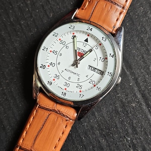 May include: A silver-toned Seiko automatic wristwatch with a white face, featuring black numerals and hands. The watch has a brown, textured leather band. The day and date are displayed at the 3 o'clock position.