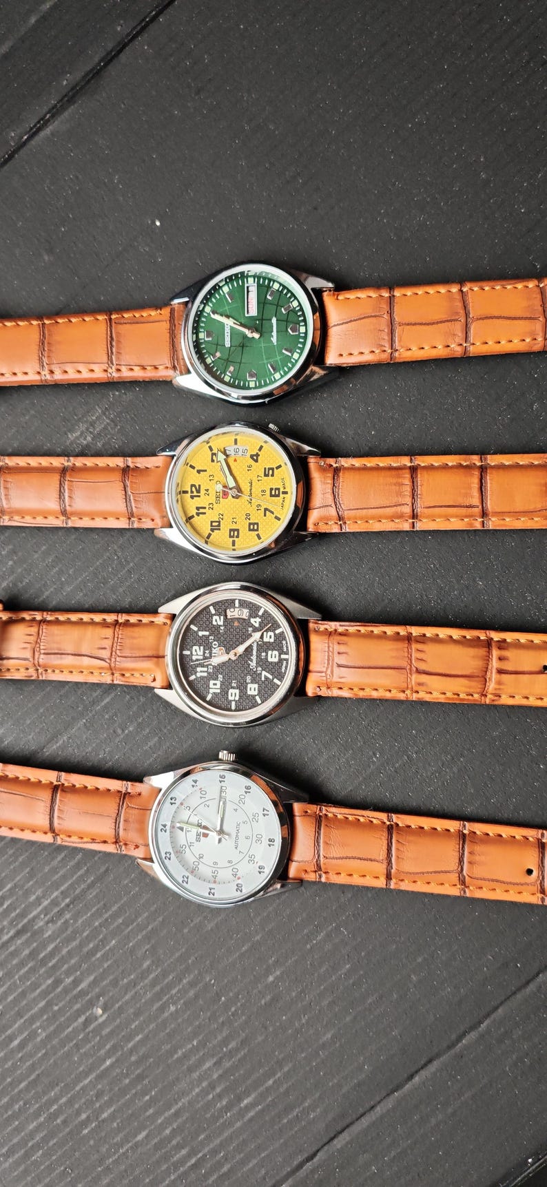 May include: Four wristwatches with brown leather straps are displayed. The watch faces are green, yellow, black, and white. The watch faces have numbers and hands. The straps have a crocodile skin pattern.