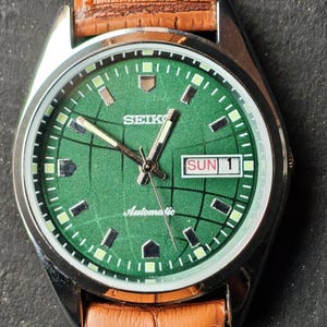 May include: A vintage Seiko automatic wristwatch with a green dial and a brown leather strap. The watch face displays the day and date, with the day reading "SUN 1". The watch has a silver-colored case.
